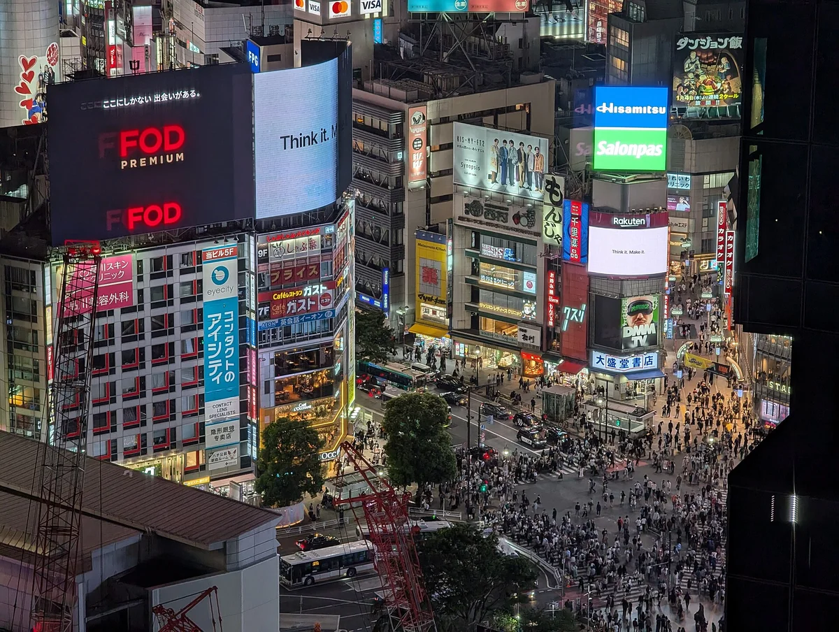 Crowds crossing the Shibuya scramble at dusk, surrounded by lit advertising screens and tall buildings