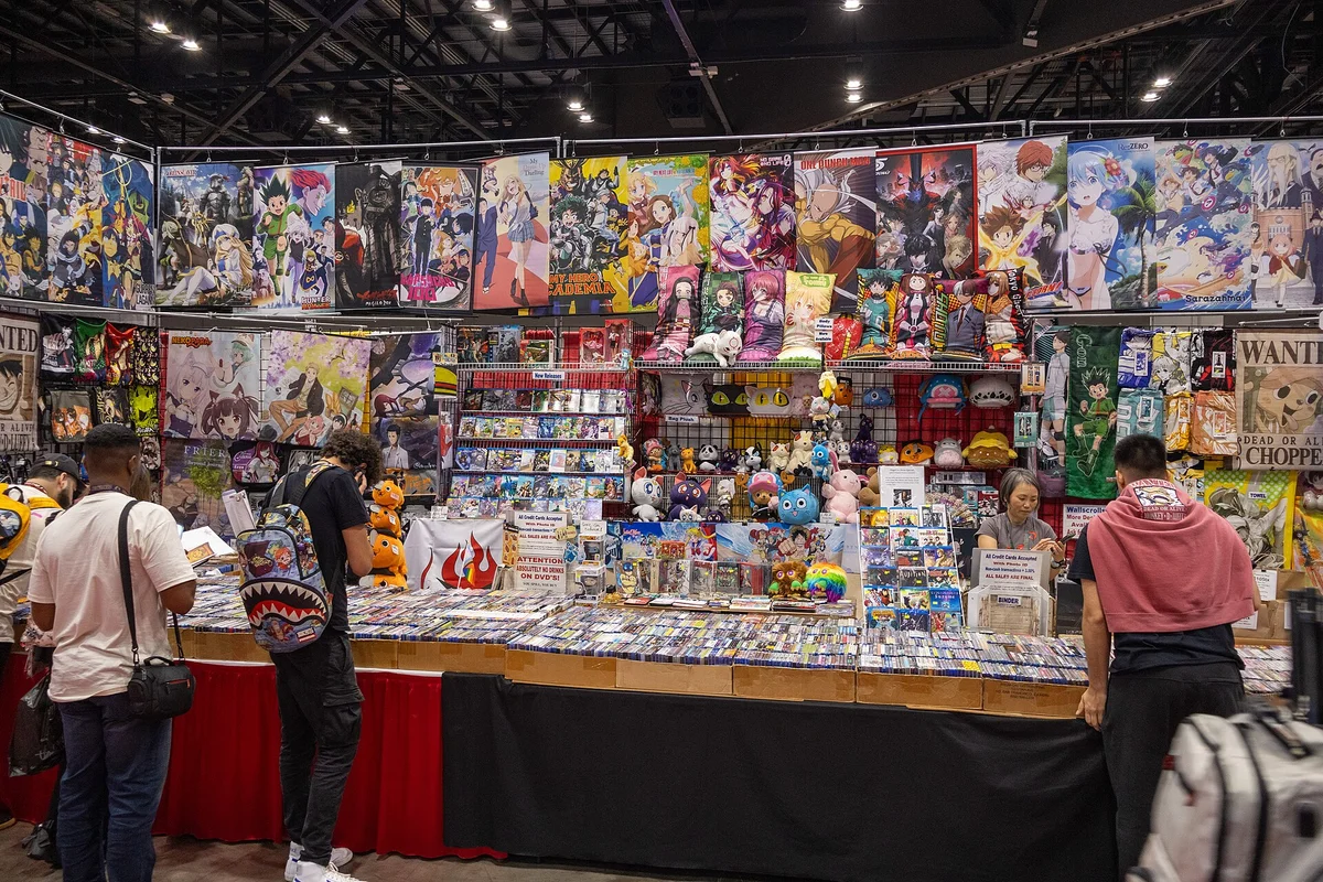 Anime convention booth with the back wall covered in rows of anime posters, shoppers browsing the tables below